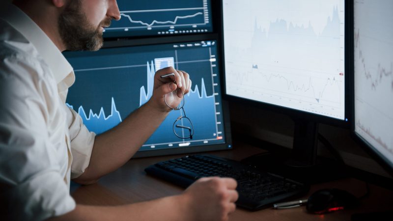 Need to grab eyewear. Bearded man in white shirt works in the office with multiple computer screens in index charts.
