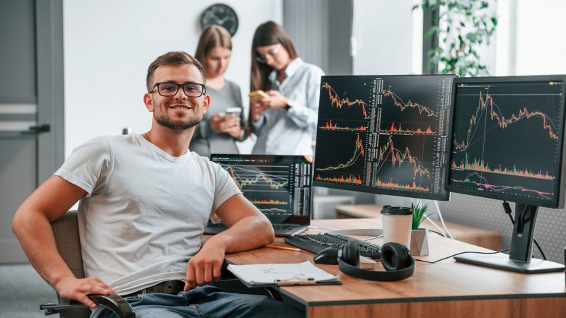 Stock broker is sitting by computer and working with exchange technology. Women is standing and talking behind.