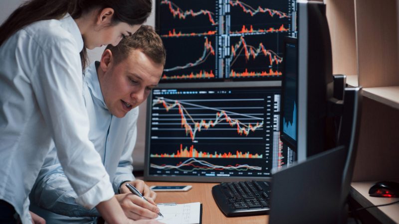 High and nice results. Team of stockbrokers are having a conversation in a office with multiple display screens.