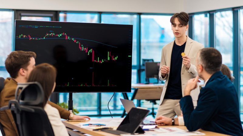 Young worker leading business meeting in an office, discussing the topic of cryptocurrencies with other workers using a big display with currency rate and physical coin